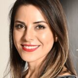 A woman with long, styled brown hair smiles warmly at the camera, wearing a striped top and bright red lipstick. The background is softly blurred, focusing on her friendly expression.