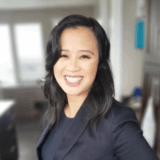 A smiling woman in a professional outfit poses for the camera, showcasing a confident demeanor. She has long, black hair and is in an office setting with windows in the background, suggesting a bright and inviting atmosphere.