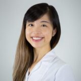 A smiling woman with long hair wearing a white lab coat poses against a light gray background, exuding a professional and approachable demeanor.