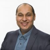 A smiling man with short hair is wearing a dark blazer over a light blue shirt. He is posed against a plain white background, exuding a friendly and approachable demeanor.