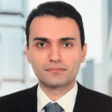 A man in a formal suit and tie poses for the camera, with a blurred cityscape background. He has dark hair and a neutral expression.