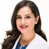 A woman with long, dark hair smiles at the camera, wearing a white coat over a blue top. She exudes a professional demeanor, likely in a healthcare or medical setting. The background is plain and light, emphasizing her friendly expression.