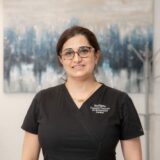 A woman in a black dental uniform stands smiling in an office setting, with abstract artwork in the background. She wears glasses and appears professional and friendly.