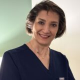 A woman in a dark blue medical uniform smiles warmly at the camera, standing against a softly lit background. Her hair is short and styled, conveying professionalism and approachability.