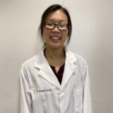 A smiling woman wearing glasses and a white lab coat stands against a neutral background. She appears to be a healthcare professional, showcasing a friendly and approachable demeanor.