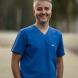 A smiling male healthcare professional stands outdoors, wearing blue scrubs. He has short, silver hair and a confident demeanor, with trees visible in the background.