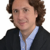 A man with wavy brown hair is smiling, wearing a black suit jacket over a blue and white striped shirt. He is facing the camera with a neutral background.