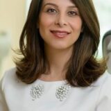 A smiling woman with long, wavy hair, wearing a white blouse adorned with pearl-like embellishments, poses in a well-lit environment. The background is softly blurred, suggesting a professional setting.