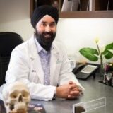 A smiling doctor wearing a white coat and a turban sits at a desk, hands clasped, with a skull model and phone on the table. Green plants are visible in the background, creating a welcoming office environment.