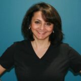 A smiling woman with shoulder-length dark hair wearing a black top stands against a teal background, posing with her hands on her hips.