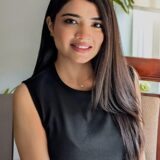 A young woman with long, dark hair sits in a chair, smiling warmly at the camera. She wears a black sleeveless top and is surrounded by a bright, airy setting with greenery in the background.