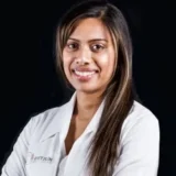 A smiling professional wearing a white lab coat poses with arms crossed against a dark background.