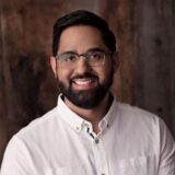 A smiling man in a white shirt and glasses stands against a rustic wooden backdrop, portraying a friendly and approachable demeanor.