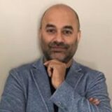 A man with a bald head and a beard is standing against a plain background, wearing a light blue blazer and a black shirt. He is thoughtfully resting his hand on his chin.