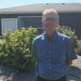 A smiling man stands outdoors in front of a gray house, surrounded by green shrubs and plants. He wears a blue shirt and appears happy in a sunny environment.