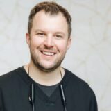 A smiling male healthcare professional wearing a dark scrub top, with a stethoscope around his neck, stands in front of a light, textured background, conveying a friendly and approachable demeanor.