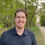 A smiling man with light brown hair and a beard stands outdoors, surrounded by trees. He wears a dark polo shirt and is positioned along a pathway, suggesting a serene, nature-filled environment.