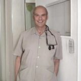 A smiling middle-aged man wearing a light-colored shirt stands near a doorway, with a pair of glasses hanging from his neck. The background shows a simple, well-lit interior.