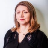 A woman with wavy blonde hair wearing a black blouse stands against a light background, looking confidently at the camera. She has a subtle smile and is wearing a necklace, conveying a professional yet approachable demeanor.