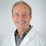 A smiling man wearing a white lab coat over a patterned shirt stands against a neutral background. He appears friendly and approachable, embodying a professional demeanor.