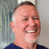 A smiling man with short gray hair and a goatee is shown. He is wearing a dark blue shirt and stands in front of a light-colored background.