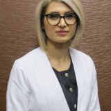 A woman with short blonde hair and black-rimmed glasses, wearing a white coat over a black top, poses against a textured brown background.