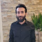 A man with a beard stands against a textured stone wall, wearing a black button-up shirt. In the background, there is a green potted plant.