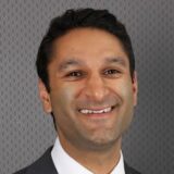 A smiling individual in professional attire, with short dark hair and a clean shaven face, set against a textured gray background.