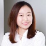A smiling woman with medium-length brown hair, wearing a white shirt, poses in front of a light gray background. Her expression is friendly and approachable.