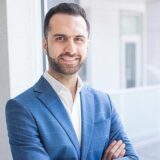 A man in a blue blazer stands confidently with his arms crossed, smiling at the camera. He has a well-groomed beard and short hair, set against a bright, modern background.
