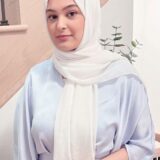A young woman in a light-colored ensemble and white hijab stands in a well-lit interior space. She has a neutral expression and poses in front of a staircase with decorative plants in the background.
