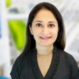 A woman with long, dark hair smiles gently, wearing a black top. The background features soft, abstract shapes in green and blue, suggesting a modern, professional environment.
