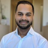 A man with a well-groomed beard smiles at the camera, wearing a white button-up shirt. The background is softly blurred, suggesting a professional setting.
