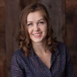 A smiling young woman with curly hair wearing a dark floral blouse is posing against a wooden background.