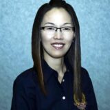 A woman with long straight hair and glasses smiles for the camera, wearing a dark shirt with a decorative emblem. She is positioned against a light blue background, giving a friendly and professional impression.