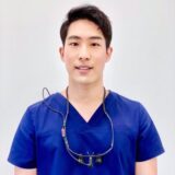A young male professional in a blue scrubs outfit is smiling for the camera, with dental loupe glasses hanging around his neck.
