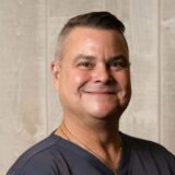 A smiling man with short hair, wearing a dark shirt, poses for a photo against a light wooden background.