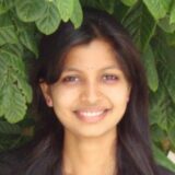 A young woman with long black hair smiles brightly against a backdrop of green leaves. She has a small nose stud and wears pink earrings. The photo conveys a warm and friendly demeanor.