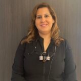 A smiling female dentist stands in a professional setting. She wears a black shirt and has dental instruments around her neck, indicating her role in dental care. The background is a simple, neutral color.