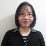 A woman with shoulder-length black hair and glasses smiles for the camera. She is wearing a black top with a striped collar and a striped shirt underneath, set against a plain light background.
