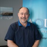 A smiling man in a blue shirt stands in front of a blue banner with the word "AQUA." The background features a soft-focus image of a beach scene.