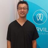 A smiling dentist wearing a black scrub top stands in front of a blue dental office sign, promoting a welcoming and professional atmosphere.