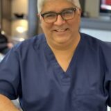 A smiling man wearing dark blue scrubs and glasses sits at a counter, appearing friendly and approachable. The background features soft lighting and blurred elements, suggesting a professional environment.