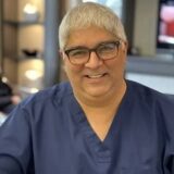 A smiling individual in blue scrubs and glasses is seated in a well-lit environment, conveying a friendly demeanor.