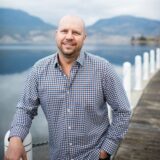 A smiling man in a checkered shirt stands by a serene body of water, with mountains in the background. He rests against a railing, exuding a relaxed and approachable demeanor.