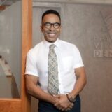 A smiling man in a white shirt and patterned tie stands in a well-lit office setting, leaning against a wooden partition. Behind him, a sign for a dental office is visible, adding context to the image.