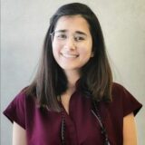 A young woman with shoulder-length dark hair wearing glasses smiles at the camera. She is dressed in a burgundy blouse and has a neutral background. Her friendly expression conveys warmth and approachability.