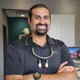 A smiling male dentist stands with arms crossed, wearing a black polo shirt and dental loupes around his neck. The background features a modern dental office setting with equipment and a computer.