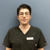 A young man with dark hair and glasses is wearing a black medical scrub top. He stands against a plain blue background, smiling confidently.