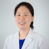 A smiling healthcare professional wearing a white lab coat and blue top, with dark hair pulled back, poses against a light background.